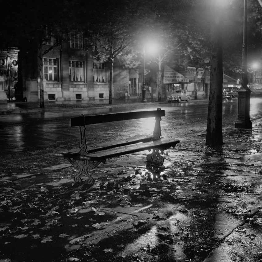 black and white photo of an empty bench on a street at night
