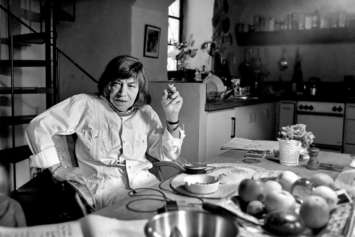 A black-and-white photo of Patricia Highsmith smoking at her kitchen table in Tegna, Switzerland